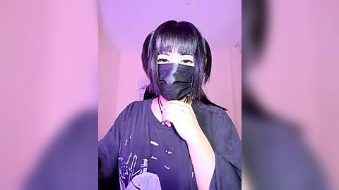 Media: A video of an East Asian woman with a pale complexion, wearing a black face mask, black t-shirt, and black headphones, against a soft pink background, with a blurred figure in the foreground.