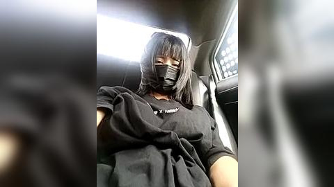 Media: A video captures a person wearing a black face mask, black T-shirt, and black gloves, sitting in a car with a blurred background.