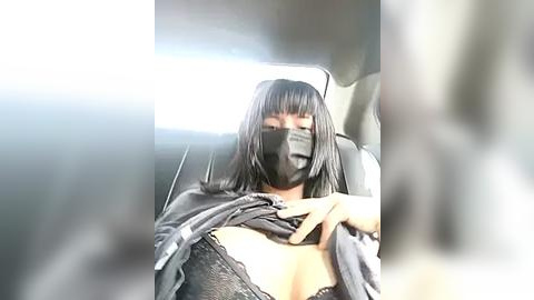 Media: Video of a woman with medium-length black hair, wearing a black face mask, black lace bra, and gray robe, sitting in a car.