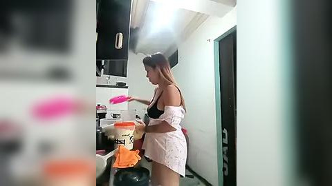 Media: A video captures a woman in a white robe, holding a pink dish, standing in a small, cluttered kitchen. The image is slightly blurred.