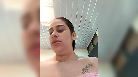 Media: Video of a Latina woman with dark hair, light skin, and a tattoo on her shoulder, wearing a pink towel, looking slightly downcast.