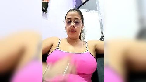 Media: Video of a Latina woman with medium skin tone and glasses, wearing a bright pink tank top with white lace trim, sitting in a brightly lit room with white walls.