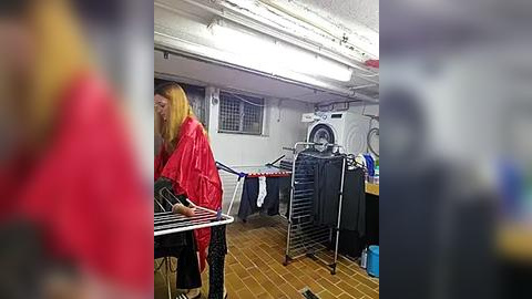 Media: Video of a woman in a bright red raincoat standing beside a clothes drying rack in a dimly lit laundry room with a washing machine and white walls.