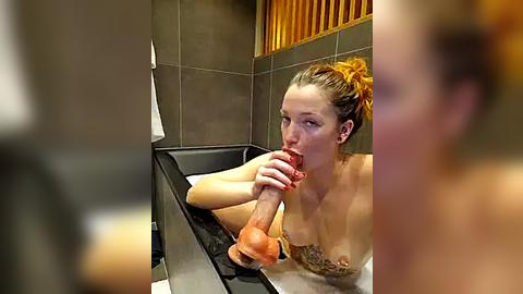 Media: Video of a nude woman with light skin and orange hair in a bathtub, holding a realistic, red dildo, with a tattoo on her left breast.