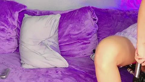 Media: A video shows a woman with fair skin, wearing a purple velvet dress, sitting on a bed covered in purple sheets. Her bare buttocks are visible as she bends forward, holding a silver object.