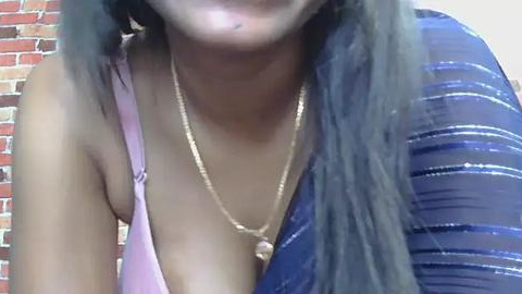 Media: Video of a woman with medium brown skin and long, straight black hair, wearing a pink bra and a blue dress with silver stripes. She has a thin gold necklace. Background shows a red brick wall.