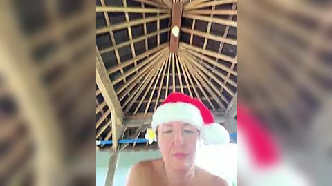 Media: A blurry video of a middle-aged woman wearing a red and white Santa hat, sitting indoors with a thatched roof and wooden beams in the background.