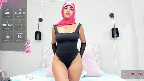 Media: Video of a woman in a black one-piece swimsuit and pink hijab, standing in a modern, white bedroom with floral decor.
