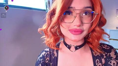 Media: A video of a woman with wavy, vibrant orange hair, wearing glasses, a black choker, and a patterned blouse, smiling, in a well-lit room.