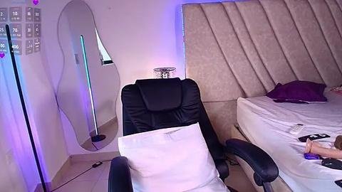 Media: Video of a modern, minimalist bedroom with a black leather chair, a large, mirrored vanity, a plush headboard, and a neatly made bed with purple pillows.