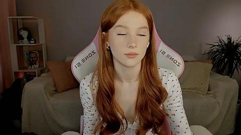 Media: Video of a pale-skinned, red-haired woman with long wavy hair, wearing a white dress, sitting on a pink and white gaming chair in a dimly lit, cozy living room.