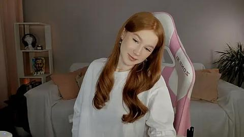 Media: Video of a fair-skinned, red-haired woman in a white sweater, seated on a pink gaming chair in a cozy, softly lit living room with a grey couch and bookshelves in the background.