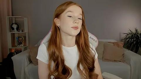 Media: Video of a fair-skinned redhead girl with long wavy hair, wearing a white t-shirt, sitting on a gaming chair in a cozy living room with beige furniture and a plant.