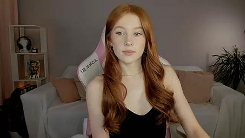 Media: Video of a young woman with long, wavy red hair, fair skin, and a slender build, seated on a pink gaming chair in a cozy, dimly lit living room with beige walls and a white sofa.