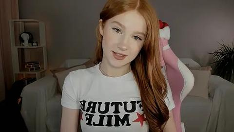 Media: Video of a young woman with long, wavy red hair, fair skin, and green eyes, wearing a white T-shirt with \"FUCK THIS\" in bold black letters. She stands in a cozy living room with light gray furniture and a pink blanket draped over her shoulder.