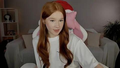 Media: Video of a young, fair-skinned redhead woman with long, wavy hair, wearing a white T-shirt, sitting on a couch in a cozy living room. Background includes a stuffed animal and bookshelves.