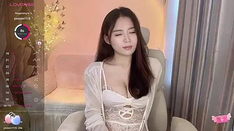 Media: A video of a young Asian woman with long dark hair, wearing white lace lingerie, sitting in a cozy living room with soft lighting and a floral background.