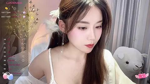 Media: Video of an East Asian woman with long brown hair, wearing a white off-shoulder top, standing in a softly lit room with white curtains and a plush teddy bear.