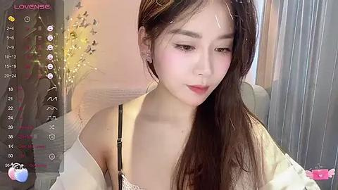 Media: A video of an Asian woman with long dark hair, wearing a white off-shoulder top, in a dimly lit room with a floral arrangement, taken with a smartphone.