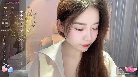 Media: A video of an East Asian woman with long, straight brown hair, wearing a white blouse, sitting indoors with a soft, blurred background.