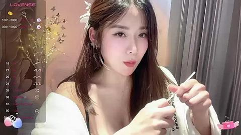 Media: Video of an East Asian woman with long, dark hair, wearing a white off-shoulder top, applying lipstick. The background features a blurred, warm-toned room with floral decor.