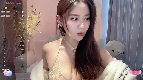 Media: Video of an East Asian woman with long brown hair, wearing a beige lace bralette, pink lipstick, and a white jacket. Background includes a soft pink wall, a teddy bear, and a vase with dried flowers.