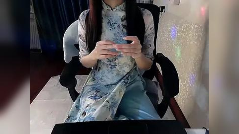 Media: Video of an Asian woman in a light blue, floral-patterned cheongsam, sitting at a desk with a laptop, wearing a black cardigan, in a dimly lit room with a white, floral-patterned wall and a red carpet.