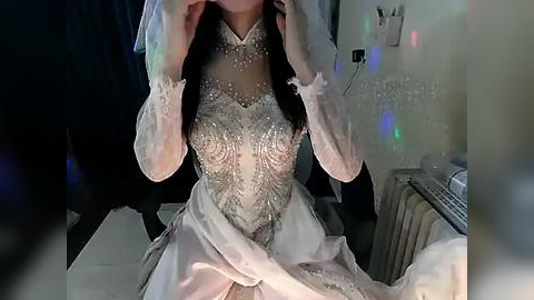 Media: Video of a woman with medium skin tone, wearing an elaborate, beaded, white wedding gown with long sleeves, seated at a desk. Background includes a dark jacket, white keyboard, and colorful holiday lights.