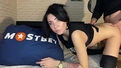Media: Video of a pale-skinned, black-haired woman in a black crop top and black thigh-high socks, bent over a bed with a blue pillow featuring the \"MOSTBET\" logo.