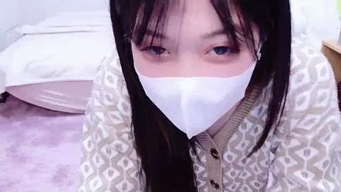 Media: Video of an Asian woman with long black hair and fair skin wearing a white surgical mask, blue eyes, white and beige patterned jacket, sitting on a pink carpeted floor in a bedroom with white bed and pillows.