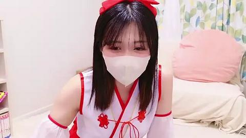 Media: Video of an East Asian woman in a white, sleeveless Chinese dress with red accents, wearing a white face mask, sitting on a bed with pink pillows in a softly lit room.