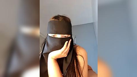 Media: Video of an Asian woman with long black hair, wearing a black mask and sleeveless top, posing against a plain grey wall.