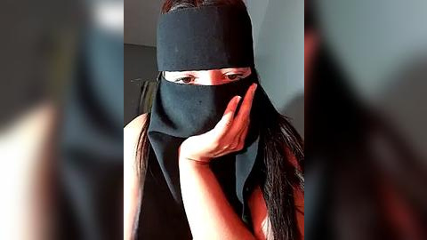 Media: Video of a woman with long dark hair wearing a black headband and mask, covering her face, holding her hand up to her mouth, in a blurred indoor setting.