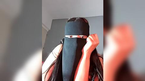 Media: A video of a person with long black hair, wearing a black face mask and a black headband, standing against a plain grey wall. The image is slightly blurred, emphasizing the mask's coverage.