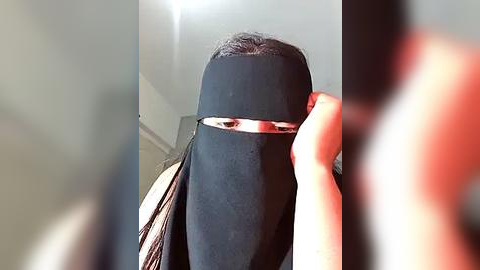 Media: A video of a person wearing a black hijab with a shiny metallic strip over their eyes, partially obscuring their face, taken from a low angle. The background is blurred, featuring a white wall and a door.