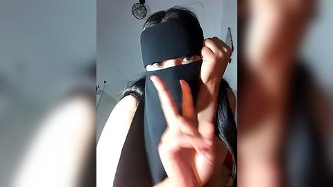 Media: Video of a person with long black hair, wearing a black face mask and black top, making a peace sign. Background is a blurred indoor setting with a clock on the wall.