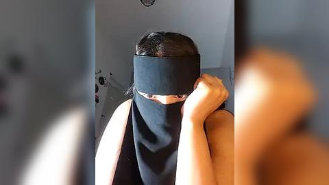 Media: Video of a woman in a black blindfold, seated indoors, holding her chin, wearing a black top, with blurred background and muted lighting.