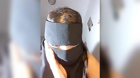 Media: Video of a person with long brown hair, wearing a black blindfold, standing indoors against a gray wall. The background is blurry, focusing attention on the blindfold.