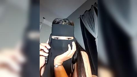 Media: Video of a woman with long dark hair, wearing a black face mask, holding a phone in a dimly lit room with a gray curtain.