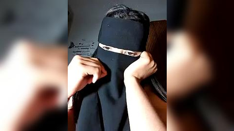 Media: Video of a person wearing a black niqab, covering their face except for their eyes, being helped to put it on by another person, blurred in the background.