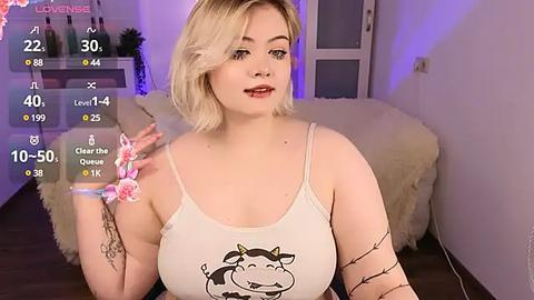 Media: Video of a fair-skinned, blonde woman with a small tattoo on her arm, wearing a cow-themed tank top, in a cozy room with soft lighting and floral decor.