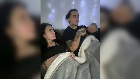 Media: A blurry video of a young woman with tattoos, wearing a black crop top, sitting next to a man with tattoos and long hair, both looking serious.