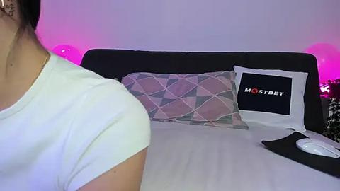 Media: Video of a woman in a white t-shirt, facing a bed with a black headboard, a patterned pillow, and a \"Moststream\" pillow. Pink and purple lights create a modern, cozy ambiance.