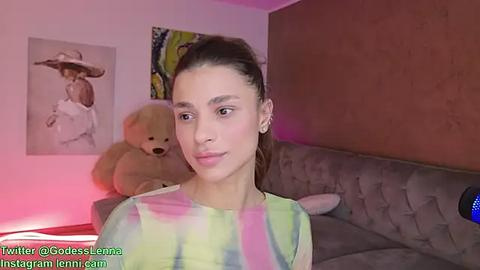 Media: Video of a young woman with dark hair, light skin, wearing a tie-dye top, in a cozy room with a stuffed bear, pink walls, and a tufted sofa.