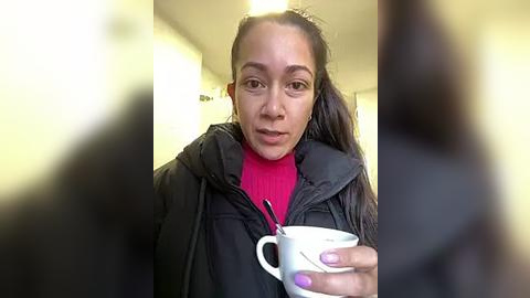 Media: A video of a young Black woman with medium skin tone and long, straightened hair, wearing a black jacket over a red turtleneck. She holds a white mug, indoors with blurred background.