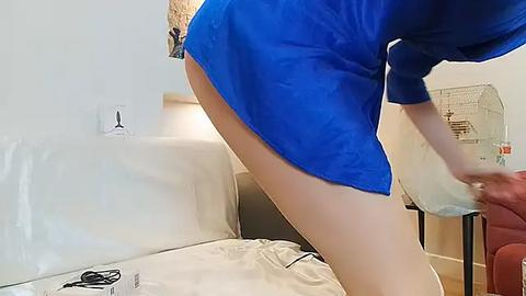 Media: A video of a person with light skin, wearing a blue robe and beige shorts, bending over a bed, holding a plastic bag, in a modern, minimalistic bedroom.