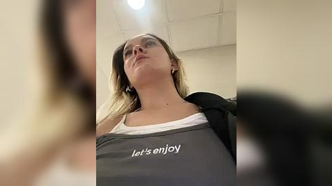 Media: A video captures a young woman with light skin and blonde hair, wearing a white tank top and gray apron with \"let's enjoy\" printed in white, standing in a dimly lit room with a white ceiling and recessed lighting.