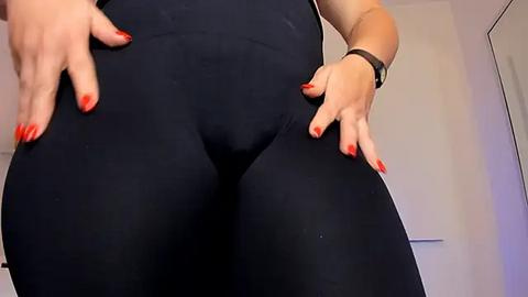 Media: A close-up video of a person with fair skin, wearing tight black leggings, red-painted nails, and a black wristwatch. The background shows a plain, light-colored wall. The focus is on the person's lower body.