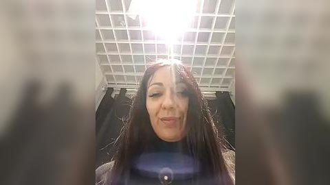 Media: Video of a smiling, fair-skinned woman with long, dark hair, wearing a dark top, taken from an upward angle in a room with a white grid ceiling.