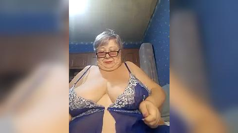 Media: Video of a plus-size, middle-aged woman with short gray hair, wearing glasses, and a blue lace bra with floral patterns, standing in a dimly lit room with blue wallpaper.